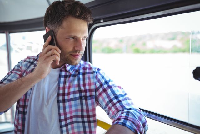 Young man in plaid shirt talking on mobile phone while traveling on bus. Ideal for use in articles about public transportation, communication, technology, and urban lifestyle. Can be used in advertisements for mobile phone services, travel apps, or casual fashion.