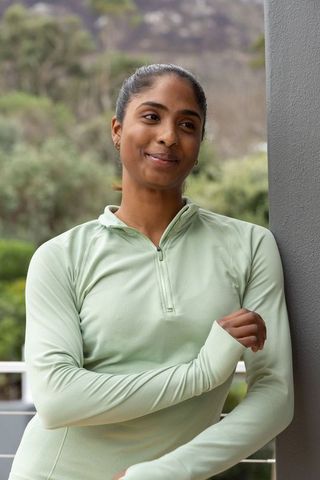 Indian woman leaning on balcony pillar wearing green zip athletic top smiling thoughtfully