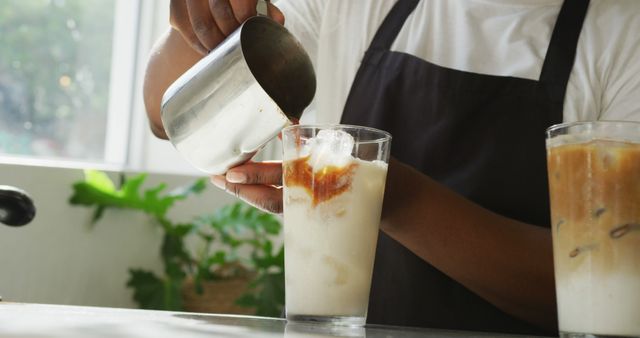 Barista pouring iced coffee into a glass with milk and ice cubes. Sunlit cafe adds a refreshing and relaxing atmosphere. Perfect for advertising coffee shops, barista work, refreshing beverages, or summer drinks promotions.