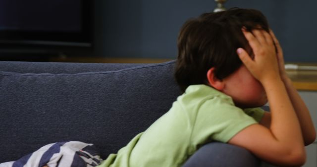 A young boy appears upset or frustrated, covering his face with his hand while sitting on a couch. His body language suggests a moment of distress or disappointment, evoking empathy and concern for his emotional state.
