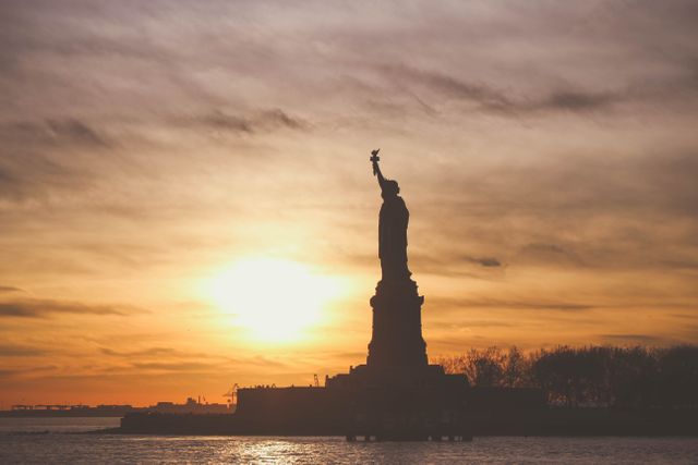 Statue of Liberty silhouette set against a vibrant golden sunset sky over water, highlighting the iconic American landmark. Ideal for use in travel brochures, tourism websites, posters, and American cultural presentations, exemplifying freedom and New York City attractions.