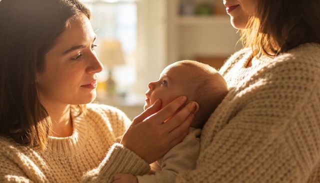 Two women cradling newborn in golden light, cozy knit sweaters, tender family moment