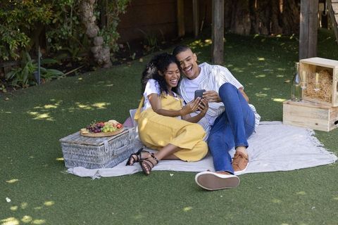 African american couple laughing on picnic blanket sharing smartphone and fruit platter