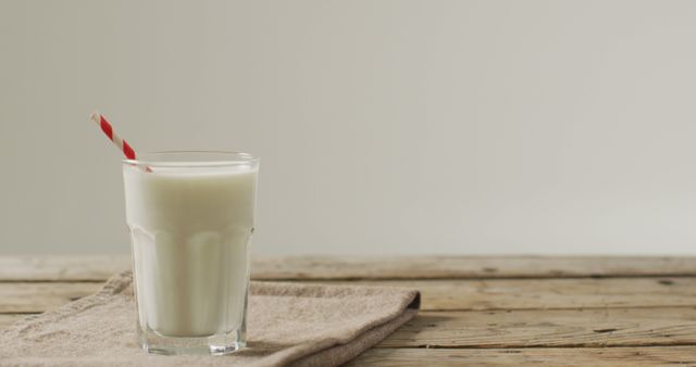 Glass of fresh milk with red striped straw placed on rustic wooden table, suggesting a natural and healthy drink. Ideal for use in dairy product advertisements, breakfast scenes, or healthy lifestyle promotions.