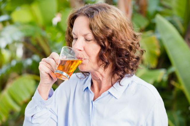 Mature woman enjoying a cup of herbal tea in a lush outdoor setting. Ideal for use in health and wellness articles, relaxation and self-care promotions, and lifestyle blogs focusing on natural living.
