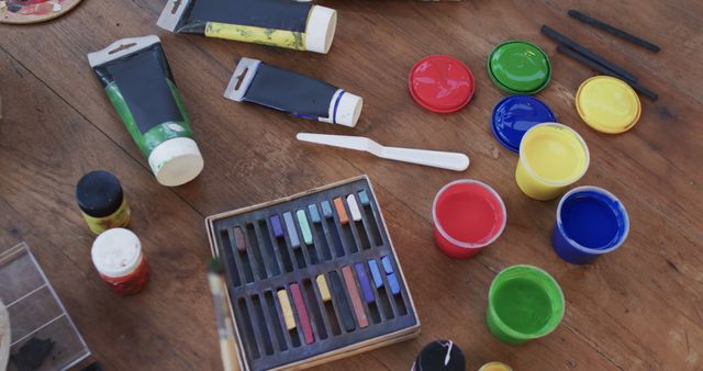 This image showcases a collection of art supplies including paint tubes, watercolor paint jars, pastels, and paintbrushes scattered on a wooden table. Ideal for use in articles or advertisements related to arts and crafts, creativity, art studios, or educational materials about artistic techniques. This can also be used to illustrate creative workshops, art classes, and hobby activities.