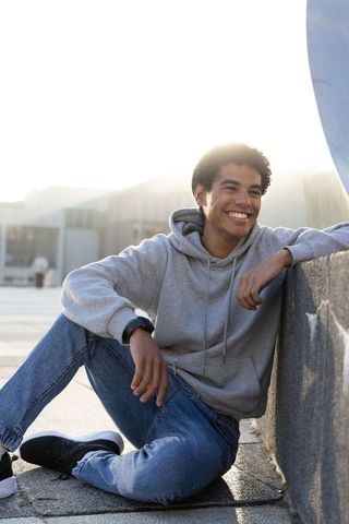 Smiling teen leaning on low wall, sitting on urban plaza wearing gray hoodie and jeans