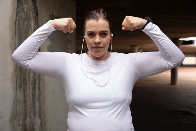 Portrait of a confident curvy Caucasian woman wearing sports clothes exercising in a city, standing in a subway flexing her biceps, smiling to camera, wearing earphones