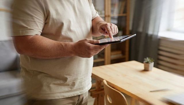 Man tapping tablet at home dining table, casual workspace with succulent and bookshelf