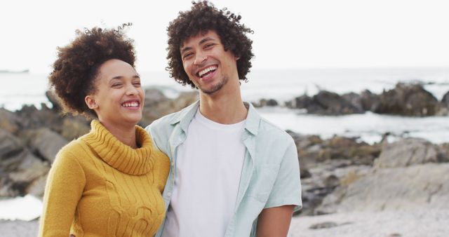 A joyful couple is enjoying a moment together by a rocky beach. Both have afro hairstyles and are dressed in casual clothing, with the woman wearing a yellow sweater. This image can be used for themes like love, happiness, relationships, and leisure. Ideal for promotional material for beach resorts, relationship counseling, lifestyle blogs, and travel brochures.