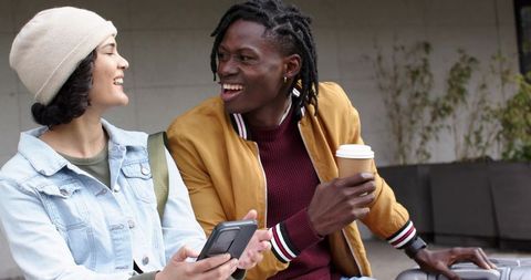 Laughing diverse friends sharing coffee and phone while sitting with luggage in urban plaza