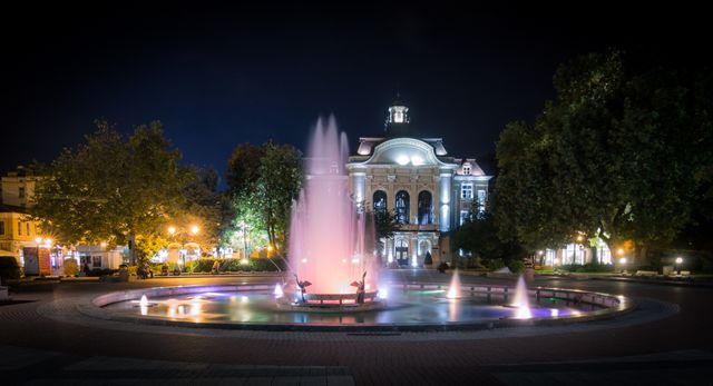 This scene captures an illuminated fountain in front of a historic building during nighttime. The vibrant lighting of both the fountain and the building creates a stunning visual effect. Ideal for use in travel blogs, websites about historic landmarks, urban cityscapes, or promotional materials for tourism in a specific city.