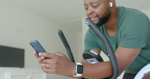 African american man checking phone while leaning on exercise bike wearing smartwatch and earbuds