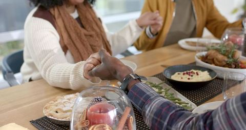 Diverse family holding hands around cozy Thanksgiving-style meal on rustic wooden table