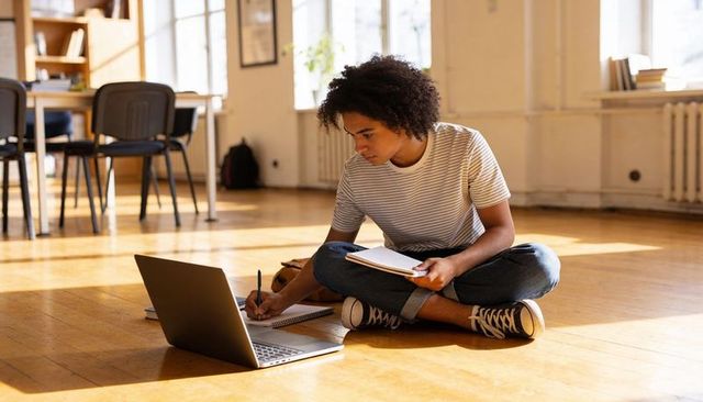 Sunlit classroom male student studying on floor, taking notes with laptop and notebook