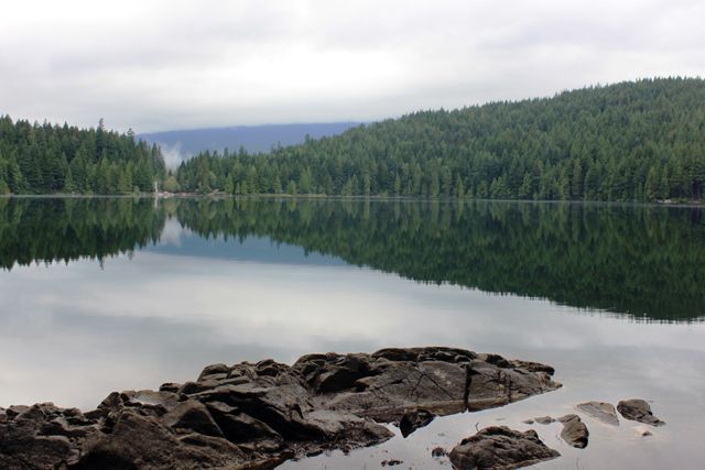 Calm lake with reflective water surrounded by dense forest creates a serene and peaceful atmosphere. Rugged rocks in the foreground add contrast to the smooth water surface. The landscape offers a tranquil retreat from everyday life, making it ideal for use in nature-related publications, travel blogs, meditation apps, and relaxation content.