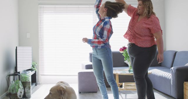 Two women in casual attire are dancing happily in a living room with abundant natural light. A dog is also visible, adding a playful atmosphere. This image can be used for lifestyle blogs, home decor advertisements, friendship concept promotions, and articles on joy and relaxation at home.