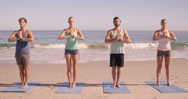 Four young Caucasian adults are practicing yoga on the beach, standing on mats in a row with a serene ocean backdrop, with copy space. Their synchronized pose and tranquil setting suggest a focus on wellness and group fitness activities.