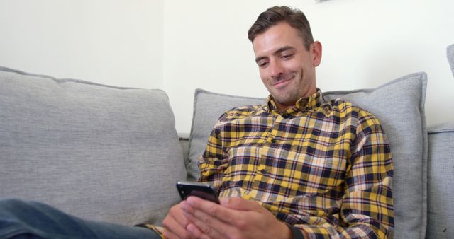 Man sitting comfortably on light gray couch, casually using smartphone with a smiling expression. Perfect for themes related to modern lifestyle, technology use, relaxation at home, and casual living. Can be used in blogs, articles, or advertisements focusing on comfort, technology, or home settings.