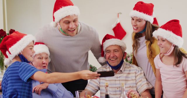 Multigenerational family celebrating Christmas, wearing Santa hats, and gathered around a table. Grandparents, parents, and children are cheerfully interacting and taking a selfie. Perfect for holiday greeting cards, promotional materials, and advertisements related to family-oriented Christmas celebrations.