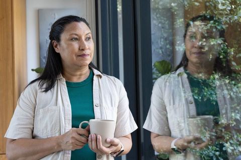 Mature african american woman standing by glass door holding mug and reflecting