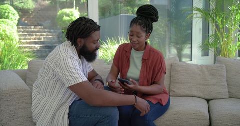 African american couple sharing smartphone while sitting on sofa in bright living room