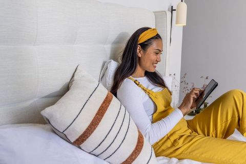 Asian woman relaxing on bed in mustard overalls using tablet in cozy neutral bedroom