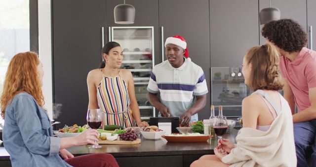 Group of diverse friends gathering in a modern kitchen, enjoying each other's company while preparing and sharing a meal. The festive Santa hat on one individual adds a holiday touch, making this perfect for content related to Christmas parties, casual entertaining, friendship, and multicultural celebrations. Suitable for blogs, social media, advertisements, and lifestyle articles showcasing joyful moments and togetherness during the holidays.