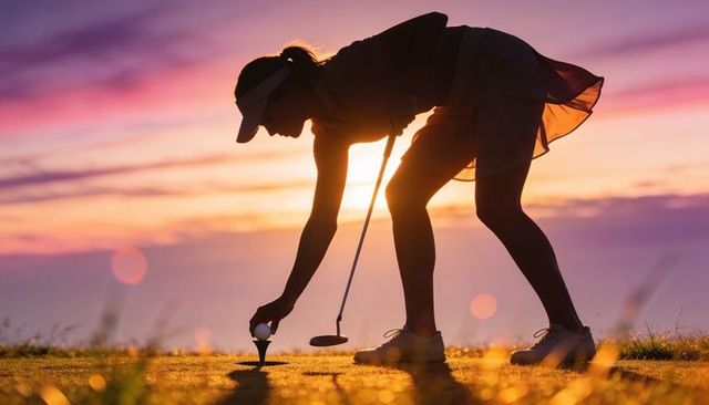Bending silhouetted female golfer placing ball on tee at golden hour sunset