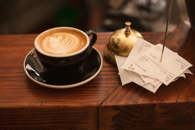 Elegant cappuccino with beautiful latte art sitting on a wooden cafe counter next to a stack of receipts and a brass bell. Useful for advertisements, cafe promotions, coffee shop websites, social media posts, and food blogs focused on cozy and elegant coffee experiences.