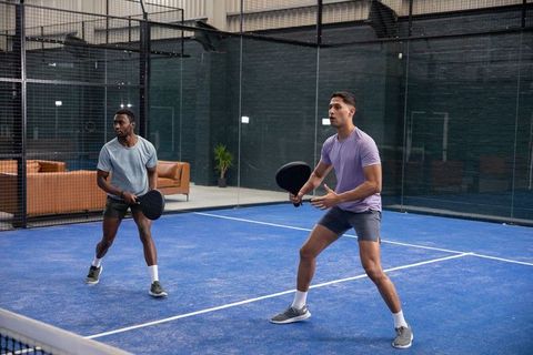Male teammates preparing to return ball on indoor blue padel court during competitive doubles play