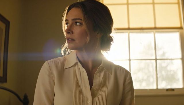 Backlit woman with bun wearing buttoned blouse gazing left in warm bedroom window flare