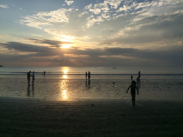 People enjoying a peaceful evening on a beach with the sun setting behind a cloudy sky. Silhouettes of adults and children create a calm and relaxing scene perfect for travel or vacation themes. This image is great for promoting tourism, vacation destinations, family outings, and connecting with nature.