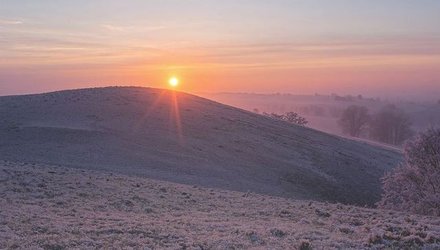 Rising sun bathing frost-covered hill with pastel sky and morning mist