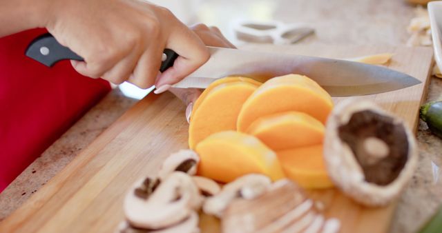 Closeup showing person slicing vegetables including orange slices and mushrooms on a wooden cutting board. Perfect for use in food blogs, recipe websites, cooking tutorials, and healthy eating campaigns. Highlights fresh, healthy ingredients and kitchen preparation.