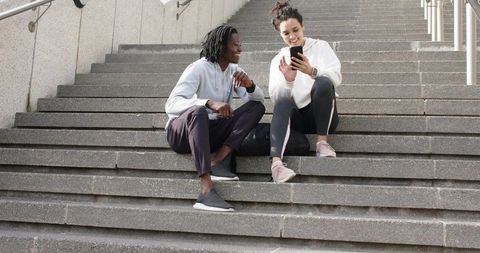 Diverse friends sitting on urban stairs sharing smartphone and laughing in sportswear