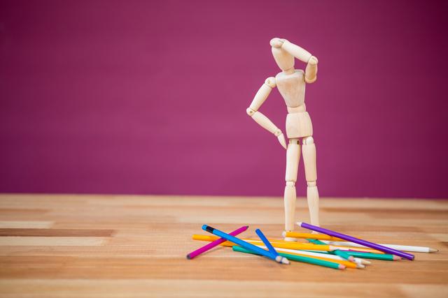 This image depicts a wooden figurine standing on a wooden table, looking at scattered colored pencils with a thoughtful pose. It is ideal for use in educational materials, creativity and art-related content, decision-making concepts, and problem-solving themes.