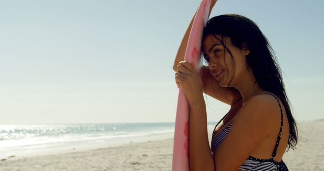 Young biracial woman holds a surfboard on a sunny beach. She enjoys the seaside ambiance, ready for a surfing adventure.