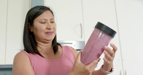 Mature woman holding purple smoothie in blender cup, inspecting healthy breakfast routine
