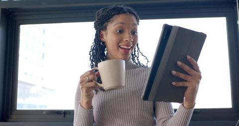 Surprised African American woman video calling on tablet while holding mug by window