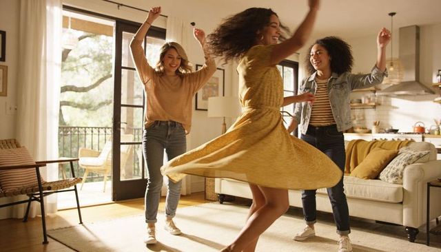 Friends dancing in sunlit living room, woman twirling in mustard yellow dress