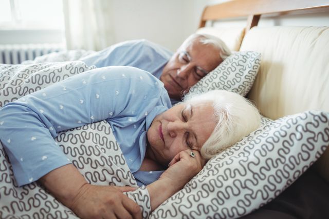 Senior couple peacefully sleeping in a cozy bedroom. Ideal for use in articles or advertisements related to senior living, health and wellness, retirement, sleep products, and family life. The image conveys themes of love, comfort, and relaxation.