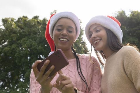 Multicultural friends wearing santa hats smiling and sharing smartphone in sunlit park