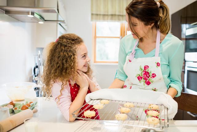 Mother and daughter smiling and interacting while baking cookies together in kitchen. Mother holding a cooling rack with cookies. Ideal for use in advertisements, blog posts, and articles about family activities, parent-child bonding, home cooking, and lifestyle.