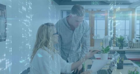 Woman guiding coworker at laptop in modern open-plan office with holographic overlay