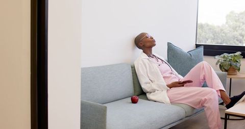 African american nurse resting on clinic sofa in pink scrubs and lab coat with tablet