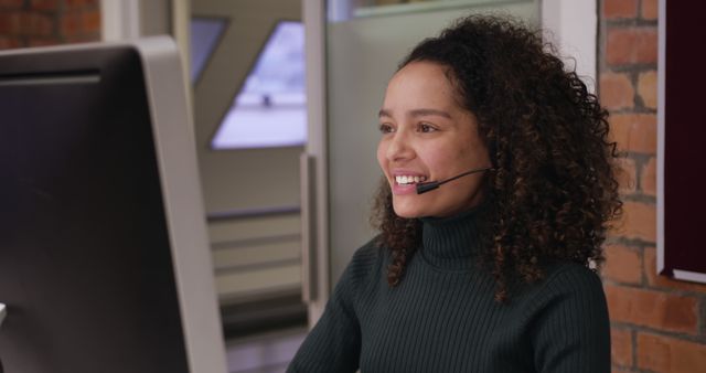 Biracial young professional wearing headset, looking at computer. She has curly brown hair, light brown skin, and is wearing dark green turtleneck