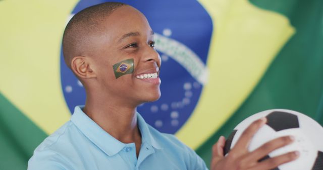 Yound boy in blue shirt smiling and holding a soccer ball, with Brazilian flag face painting, celebrating sports. Colorful background with Brazil's national flag creates festive atmosphere. This image can be used for content related to sports enthusiasm, Brazil national pride, soccer events like World Cup, and fan support.