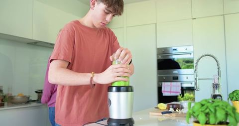 Teen male blending green basil lemon smoothie at modern kitchen island, healthy drink
