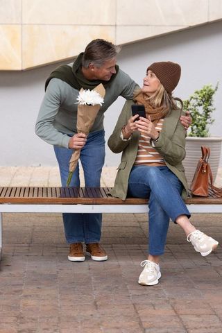 Middle-aged man offering bouquet while woman checking phone on bench in urban courtyard